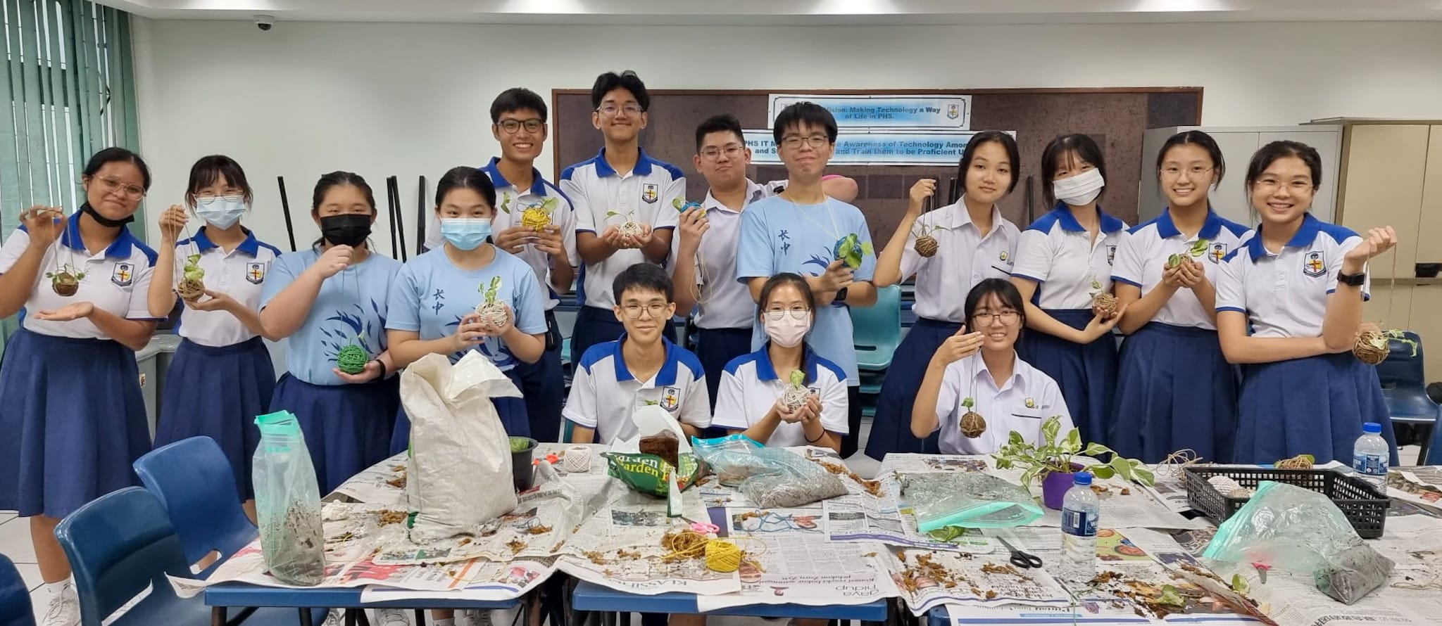 Students in school uniforms hold handmade kokedama moss balls in a classroom with craft supplies on a newspaper-covered table.